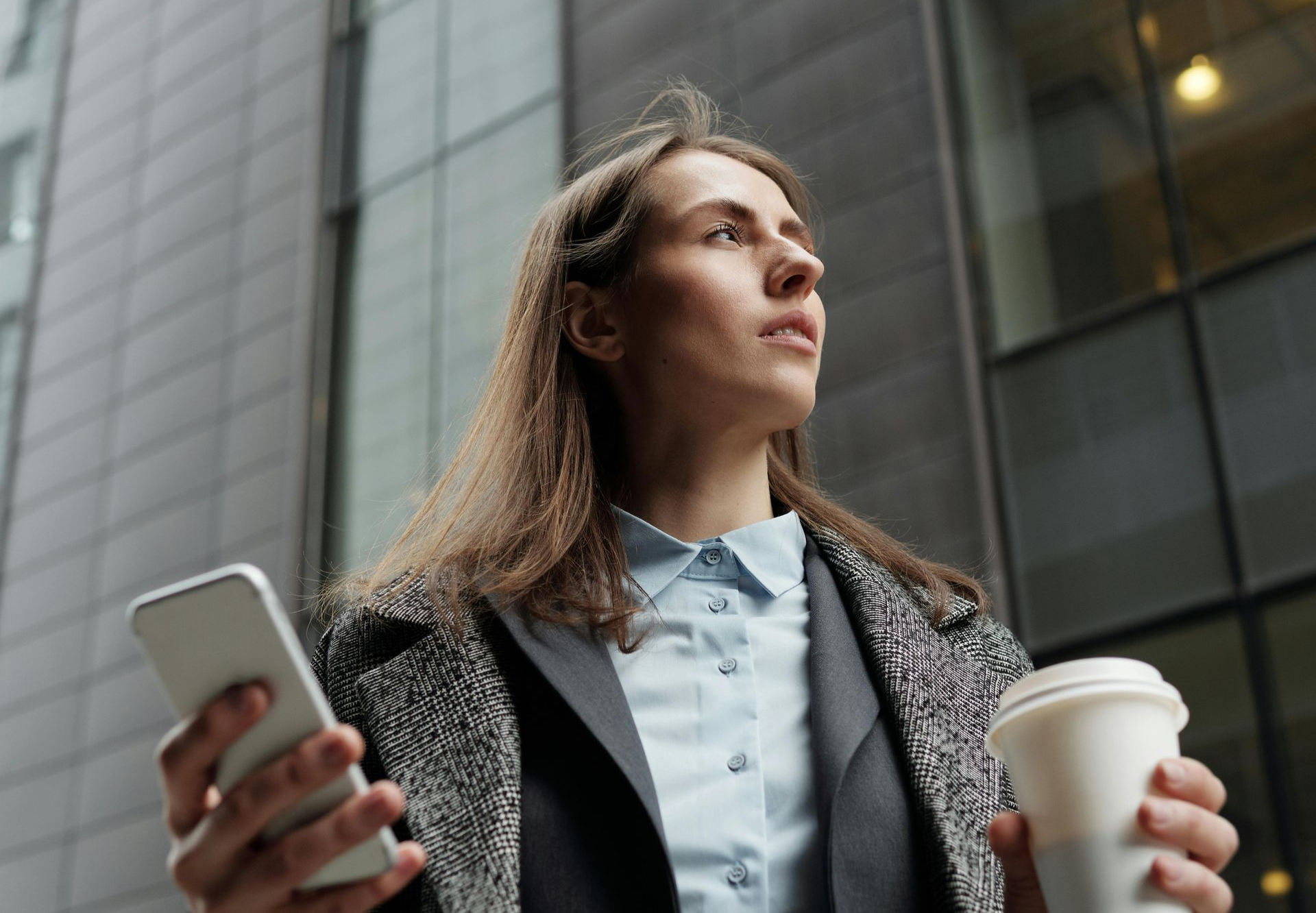 Mulher vestida com roupa de negócios segura o celular e uma xícara de café, olhando para cima perto de um prédio moderno.